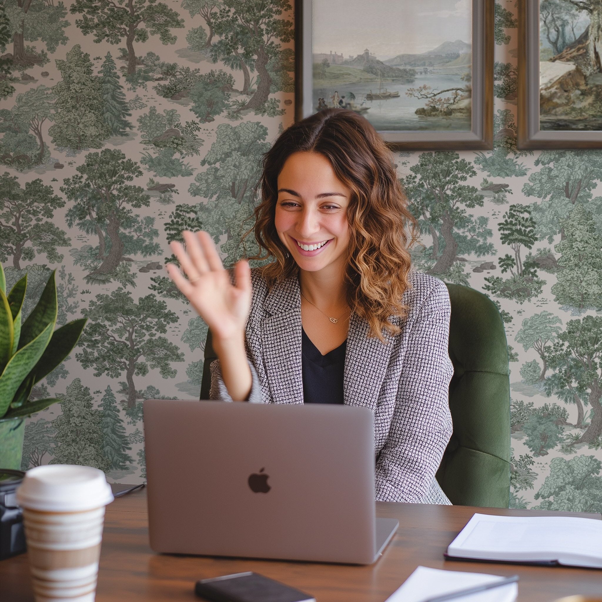 Woman waving at a laptop in an office setting with decorative wall paper.
