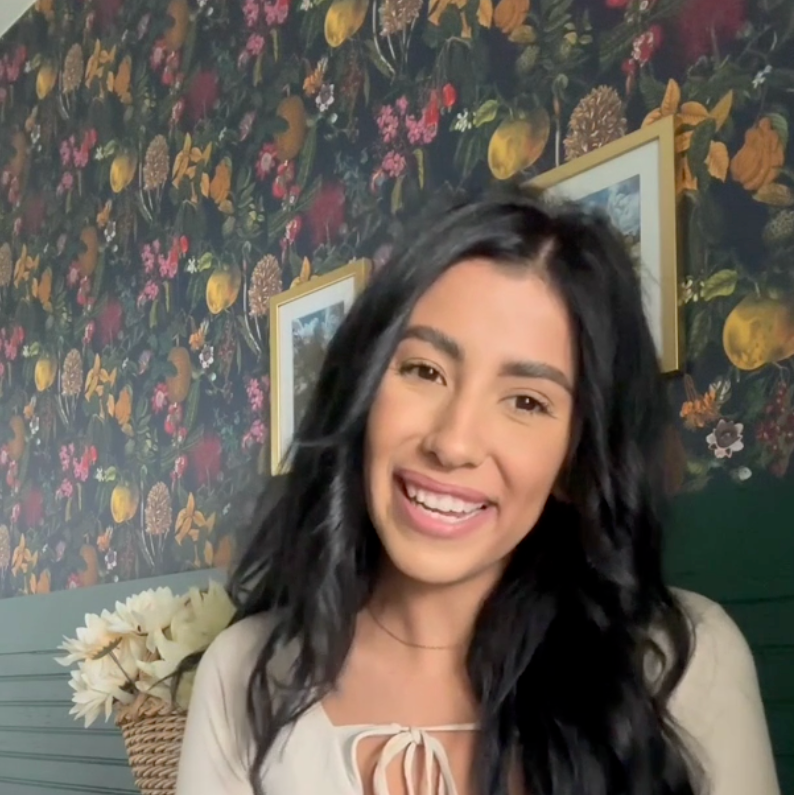 Woman with long dark hair smiling in front of a floral wallpapered wall.