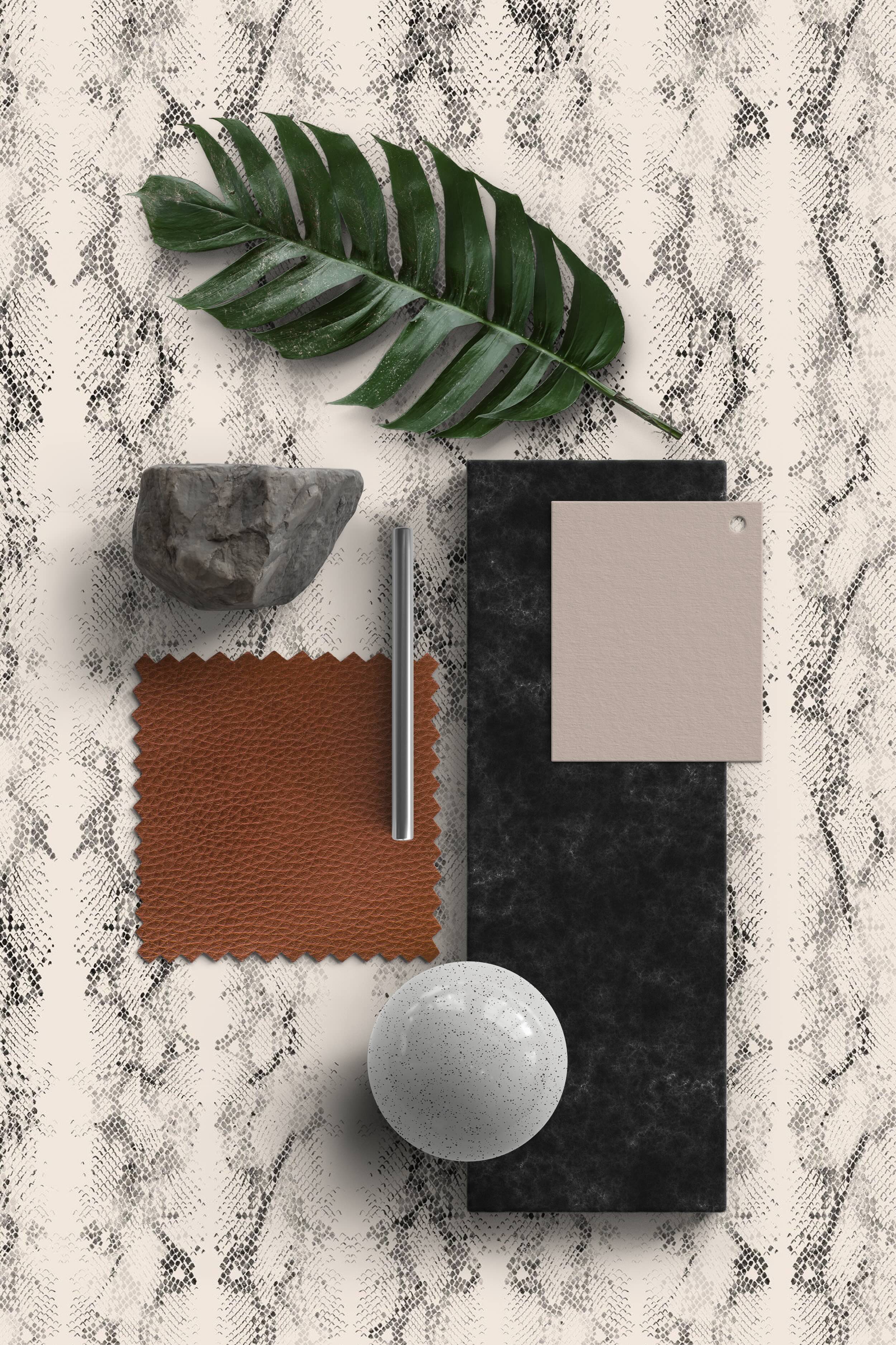 A moodboard arranged on a cream snakeskin-patterned wallpaper, featuring a large green leaf, a textured stone piece, a brown leather swatch, a metal rod, a black rectangular surface, a small beige sample card, and a speckled white sphere