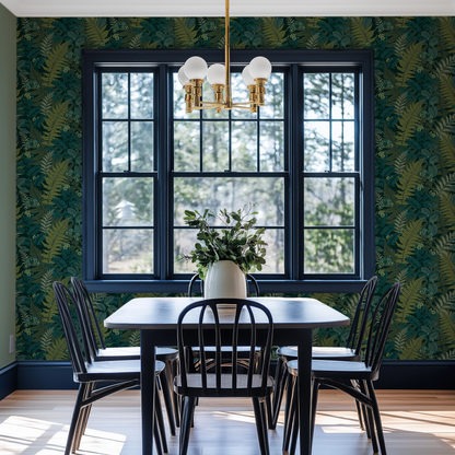 A dining room with dark blue dining table and a gold light. The wall is covered with a dark green fern leaf wallpaper and there is a bright window in the middle