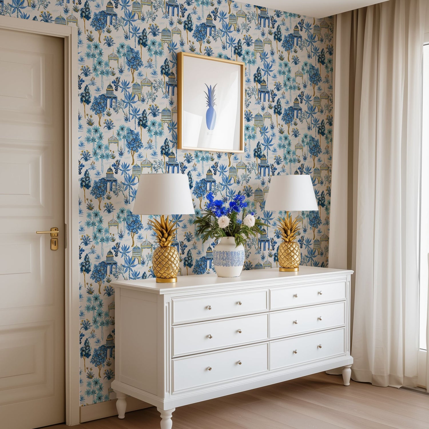 A white living room dresser with two gold pineapple lamps and white Bali Jungle wallpaper